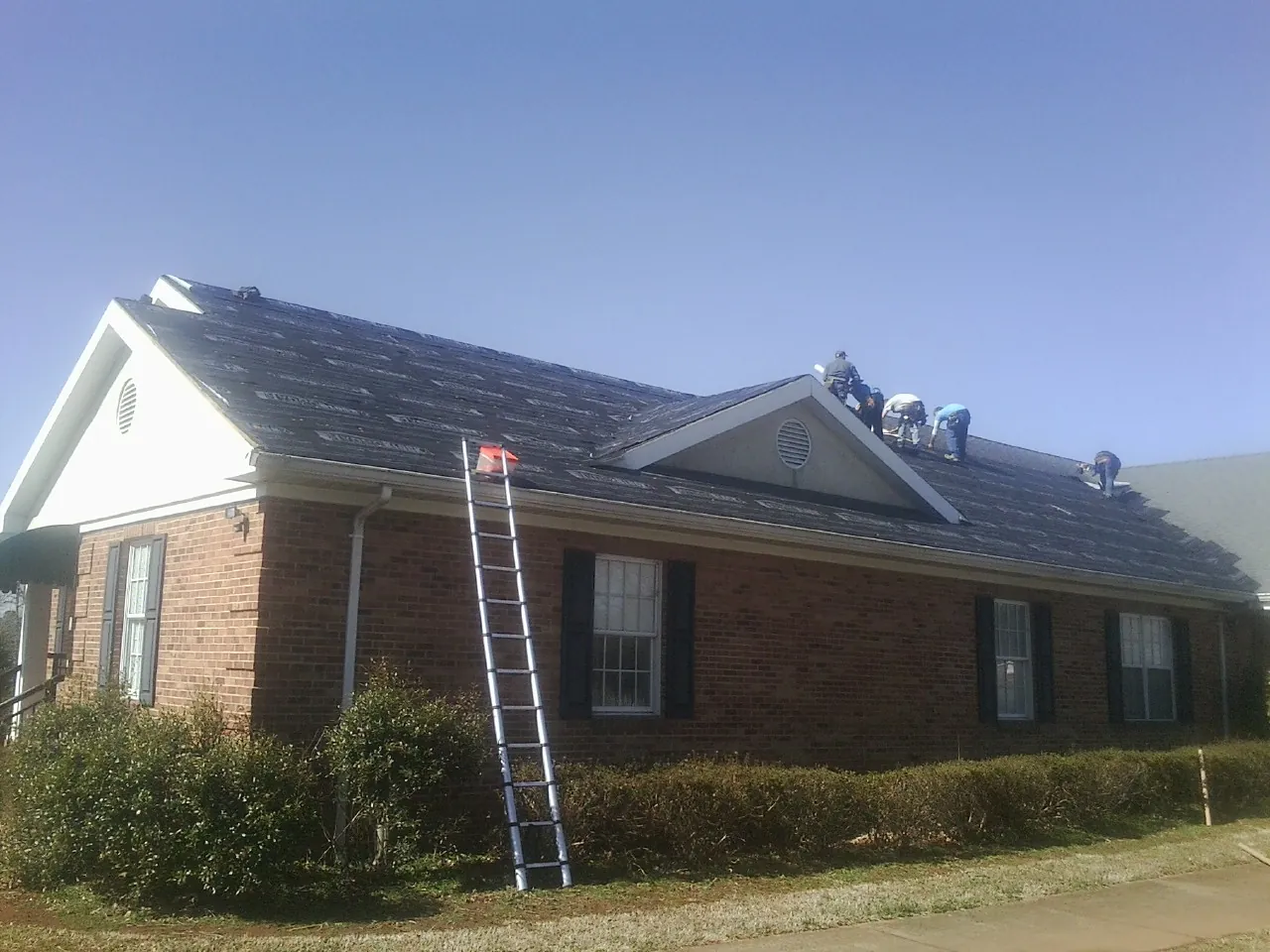 Crew tearing shingles off a brick ranch roof with a ladder set — Emergency Roof Repair work by our crew in Sutton